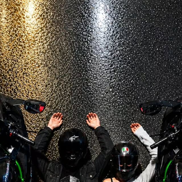 Two riders in helmets, hands raised against a glittering wall, between two Kawasaki H2s