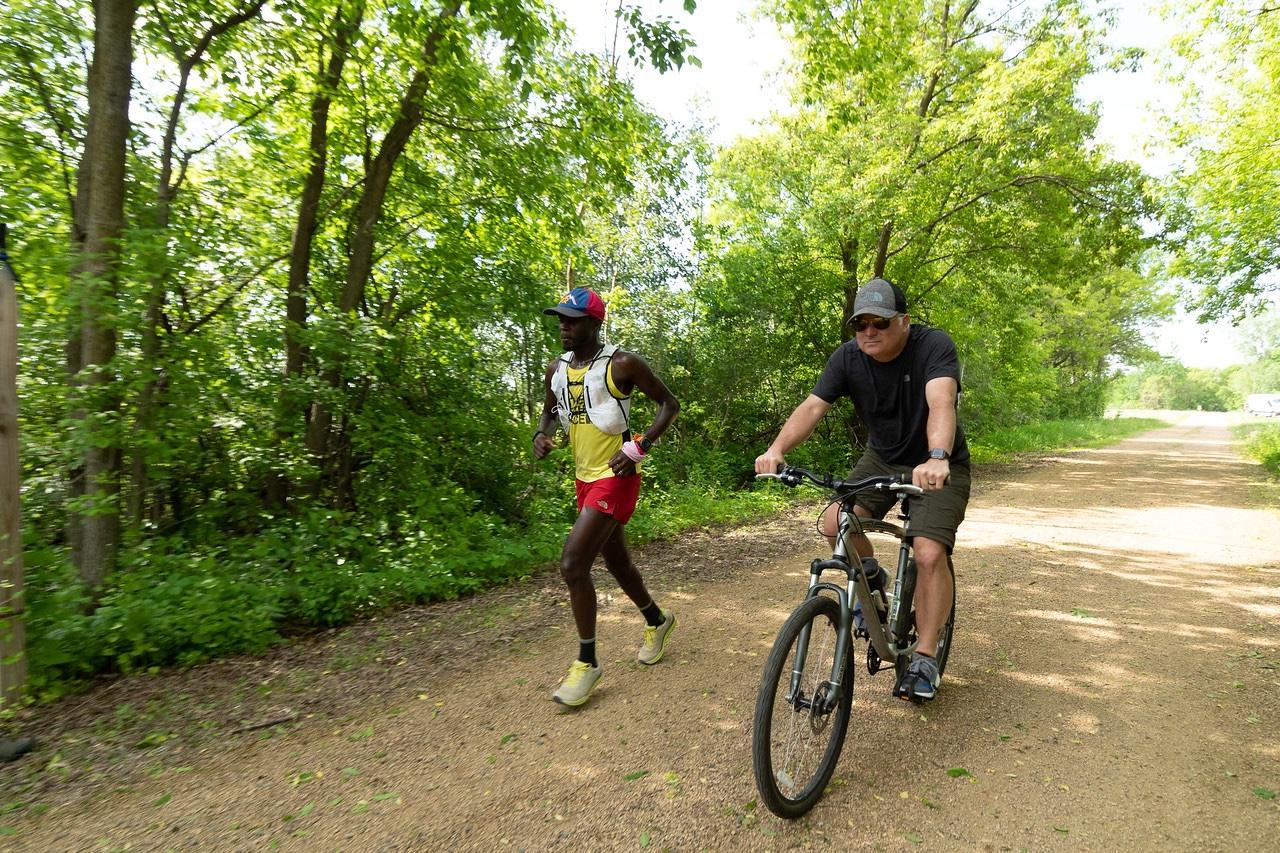 Coree running a gravel trail beside a crew member on a bike